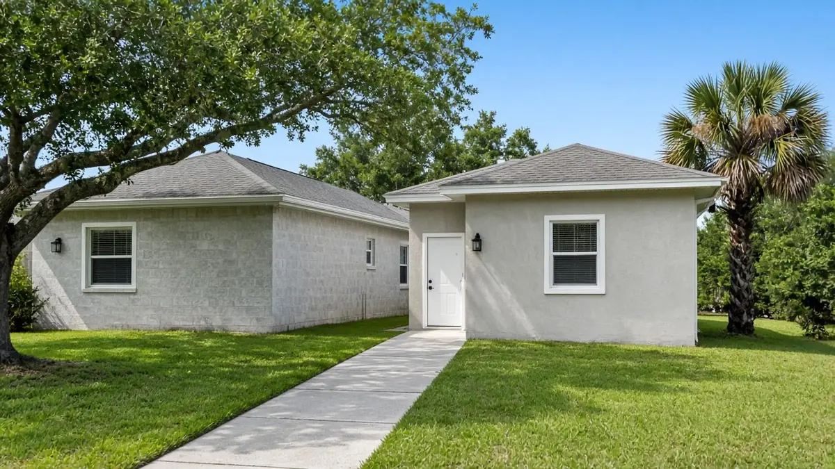 Small stucco house with a white front door and a hip roof at the end of a concrete walk, beside a larger concrete-block house, on a green lawn with an oak and a sabal palm